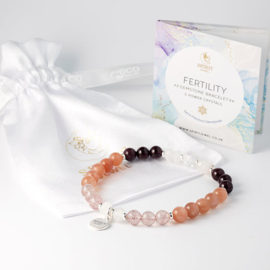 a fertility bracelet with a variety of colored beads, including orange, brown, and clear beads, arranged in a bracelet-like design. The bracelet is placed on a white surface next to a white bag with a gold logo. In the background, there is a package of fertility jewelry, which includes a booklet and a small charm.