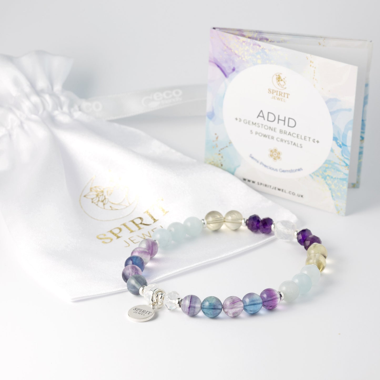 a bracelet with various colored gemstones, including blue, purple, and yellow, arranged in a row. The bracelet is placed on a white surface next to a white drawstring bag and a box with a label that reads "ADHD GEMSTONE BRACELET + 5 POWER CRYSTALS".