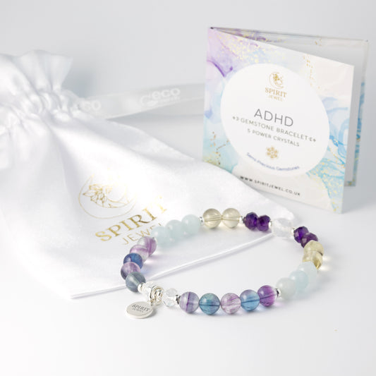 a bracelet with various colored gemstones, including blue, purple, and yellow, arranged in a row. The bracelet is placed on a white surface next to a white drawstring bag and a box with a label that reads "ADHD GEMSTONE BRACELET + 5 POWER CRYSTALS".