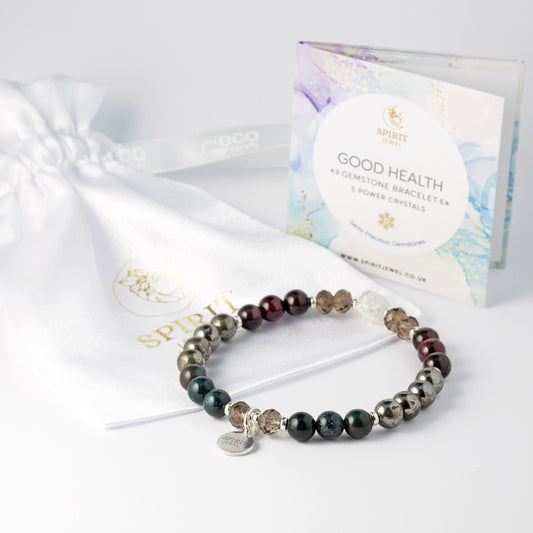 A bracelet made of colorful beads, including green, brown, and silver, rests on a white surface next to a white drawstring bag and a package of the bracelet.