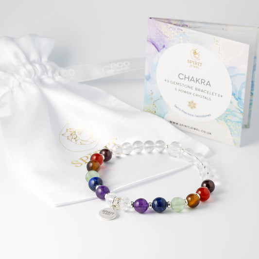 A clear crystal bracelet with colorful beads and a charm, placed next to a white gift bag with a label that reads "CHAKRA" and "Spirit Jewelry".