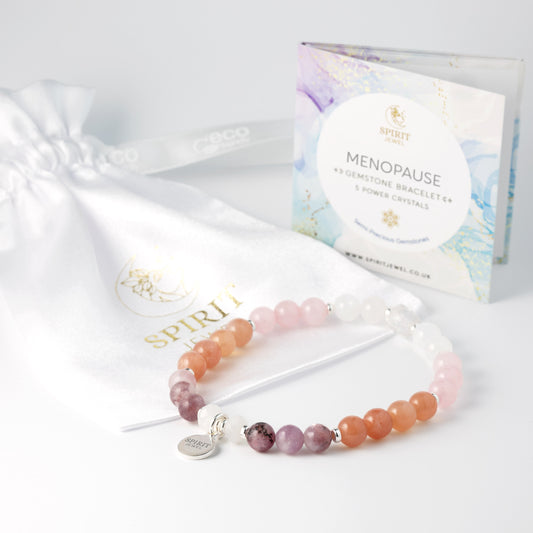 a bracelet with a variety of colored beads, including white, pink, orange, and purple, arranged in a row. The bracelet is placed next to a white bag with a gold logo and a booklet that appears to be a gift set.