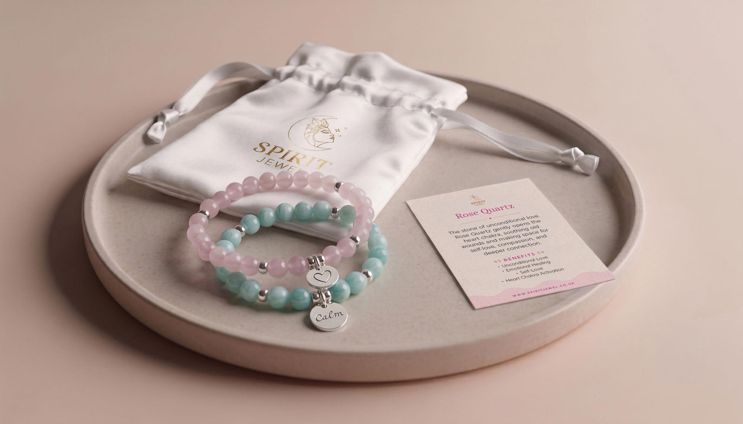An Amazonite and Rose Quartz beaded bracelet crystal set displayed on a round tray with a white drawstring pouch and a small printed card.