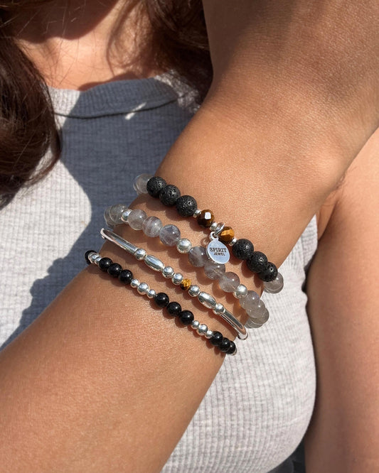 A person's arm wearing multiple bracelets, including a black and silver bracelet, a silver bracelet with black beads, and a bracelet with gray beads and a charm.
