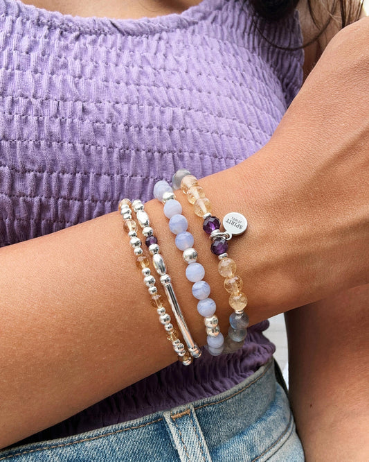 A person wearing a purple top and blue jeans is wearing multiple bracelets on their wrist.